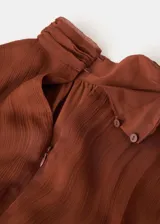 Vestido midi marrón de viscosa transparente, con diseño plisado, cuello alto fruncido y mangas largas abullonadas con puños elásticos. Presenta detalles de costuras fruncidas en la cintura y falda escalonada con vuelo.