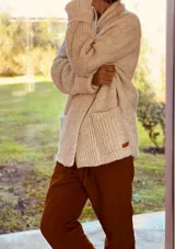 Cárdigan largo color crudo, de tejido de punto grueso con cuello alto, manga larga y bolsillos delanteros.