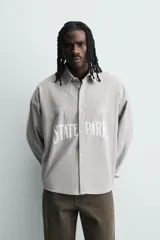Camisa de corte boxy fit confeccionada en tejido de algodón con diseño de rayas verticales. Presenta cuello solapa, manga larga con puños abotonados y cierre frontal de botones. El frente destaca por bordados y estampados a contraste.