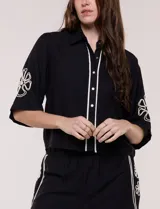 Conjunto de dos piezas negro compuesto por camisa de manga corta con cuello camisero y pantalón palazzo de tiro alto. Presenta detalles de bordados florales en contraste a lo largo de los laterales del pantalón y en las mangas.