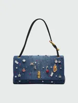 Cartera tipo shoulder bag de Marc Jacobs confeccionada en denim azul, decorada con múltiples charms metálicos y de cristal, incluyendo figuras de labios, cerezas, estrellas y dados. Cuenta con cierre frontal metálico y correas de hombro de cuero negro.