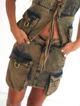Minifalda de denim color marrón ahumado con efecto desgastado. Presenta múltiples bolsillos tipo cargo con herrajes metálicos color bronce y cierre frontal con cordones.
