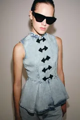Chaleco de denim claro con efecto lavado, cuello mao, sin mangas y corte peplum con pliegues en la parte inferior. Cierre frontal con botones de alamares negros.