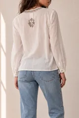 Blusa blanca de manga larga con detalles bordados en tono marrón en el pecho y las mangas, cuello redondo y cierre frontal con pequeños botones.