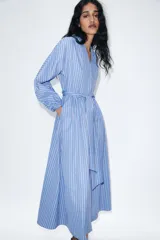 Vestido midi de corte camisero con estampado de rayas verticales en tonos azul, blanco y rojo. Presenta cuello redondo con escote en pico, mangas largas abullonadas con puños elásticos y cinturón ajustable del mismo tejido. Incluye bolsillos laterales ocultos y cierre frontal cruzado con botones.