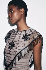 Capa o chal triangular de abalorios azul oscuro, con diseño de telaraña y tres motivos florales o de hojas bordados. El borde inferior tiene flecos largos hechos de abalorios.