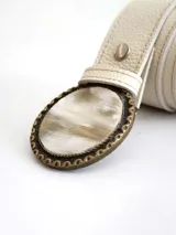 Cinto de cuero color crudo con forro de cuero y sistema de broche suizo intercambiable. Presenta una hebilla circular de guampa con borde metálico labrado en tono bronce.