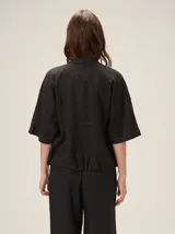 Chaqueta de lino y rayón color tostado, de corte holgado, mangas cortas amplias, cuello mao y cierre frontal oculto con cremallera. Presenta pespuntes a contratono en las costuras y cordón ajustable en el bajo.