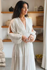 Vestido largo blanco de estilo bohemio, con escote en V profundo, mangas largas abullonadas y cintura fruncida elastizada. Presenta un diseño fluido con sutiles rayas verticales tejidas en la tela y una abertura lateral en la falda.