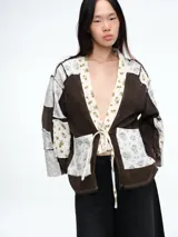 Camisa de estilo kimono con diseño patchwork, confeccionada en una combinación de tela texturizada color marrón y paneles de tela estampada con motivos florales en tonos claros. Presenta cierre frontal mediante lazo, mangas largas y bolsillos frontales.