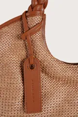 Bolso tipo shopper de textura tejida en color natural, con asas superiores trenzadas en tono marrón y detalles de ribetes en cuero sintético. Incluye un colgante decorativo de cuero con logo grabado.