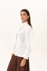 Camisa blanca de cuello alto con volados frontales a lo largo de la botonadura, manga larga con puños con volados y cierre frontal con botones.