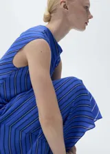 Vestido largo sin mangas con cuello drapeado y diseño evasé. Presenta un estampado de rayas diagonales en tonos azules y blancos, con cierre de botones en la parte posterior.