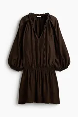 Vestido túnica corto de color marrón con leve brillo, corte oversized, cuello redondo con ribete trenzado y escote en V con tiras finas. Cuenta con mangas globo largas de corte raglán con abertura en los hombros, puños elásticos y cintura fruncida con vuelo en la falda.