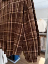 Camisa de manga larga con diseño de cuadros en tonos marrones y beige. Presenta cuello clásico, cierre frontal con botones y mangas con puños fruncidos.