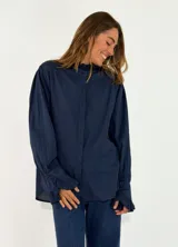 Camisa de algodón color azul marino, con cuello alto tipo mao con volados, cierre frontal oculto y mangas largas con puños elásticos decorados con volados.