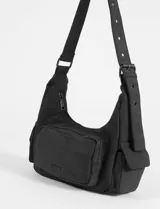 Cartera de hombro de símil gamuza color negro, con diseño de media luna, bolsillo frontal con cierre y bolsillos laterales. Cuenta con correa ajustable con ojales metálicos.