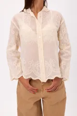 Camisa de encaje color crema con diseño floral bordado, cuello camisero, cierre frontal con botones y mangas largas con terminación acampanada.