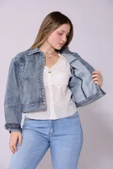 Campera de jean de mujer, color azul claro lavado, con corte cropped y estilo clásico. Presenta cuello camisero, botones metálicos frontales y bolsillos de parche en el pecho.