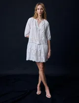 Vestido corto blanco de algodón con bordado calado tipo broderie, cuello redondo, escote en V con lazo y borlas, y mangas tres cuartos con puño.