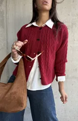 Cardigan de punto con textura trenzada en color bordó, con cuello redondo y cierre frontal mediante botones tipo alamar. Presenta mangas largas con puños acanalados y un corte clásico.