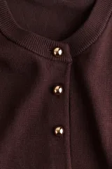 Cárdigan de punto color borgoña oscuro, con cuello redondo y manga larga. Presenta cierre frontal con botones dorados metálicos y remates acanalados en cuello, puños y bajo.