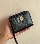 Billetera pequeña negra de cuero con textura simil lagarto, cierre perimetral y aplique metálico circular dorado con diseño grabado.