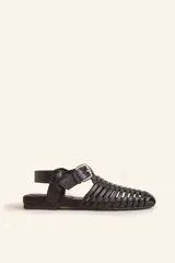 Sandalia de cuero negro estilo huarache, con puntera cerrada mediante tiras finas entretejidas a mano, tira ajustable al tobillo con hebilla metálica y suela plana con taco de 1 cm.