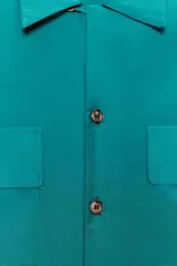Camisa de corte fluido con cuello solapa y manga larga. Presenta cierre frontal con botones y dos bolsillos de plastrón con solapa en el pecho. El diseño cuenta con bajo redondeado y costuras verticales decorativas.