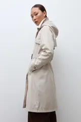 Trench impermeable color beige de corte clásico, con cierre frontal de doble botonadura, solapas amplias y cinturón ajustable con hebilla. Incluye capucha desmontable y bolsillos laterales.