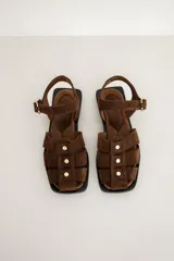 Sandalias planas de gamuza marrón, estilo pescador, con tiras entrelazadas, tachas doradas y tira ajustable con hebilla en el tobillo.