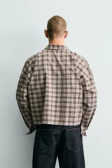 Camisa polo de corte boxy fit confeccionada en tejido de algodón con elasticidad, presenta un estampado de cuadros en tonos marrones, azules y blancos. Cuenta con cuello solapa, cierre frontal de botones, manga larga con puños abotonados y bajo con aberturas laterales.