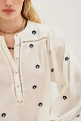 Camisa de algodón color crudo con bordados florales pequeños en azul marino. Presenta mangas largas abullonadas, cuello redondo y una abertura frontal con botones y detalles de encaje calado en la pechera y hombros.