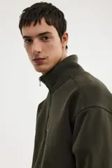 Buzo de tejido pesado de mezcla de algodón con interior cepillado suave, color verde oliva. Modelo con hombros caídos, cuello elevado con medio cierre metálico y puños y bajo en punto acanalado.