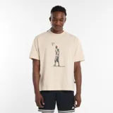 Remera de manga corta color beige claro con cuello redondo. Presenta un estampado gráfico en el centro que simula un dibujo a lápiz de un jugador de baloncesto realizando un tiro al aro, con la marca New Balance escrita debajo.