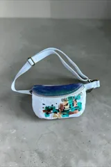 Riñonera blanca con solapa de cuero tornasolado, cierre frontal superior y correa ajustable. Presenta detalles de arte pintado a mano en el frente con inscripciones y manchas abstractas en tonos verdes, marrones y negros.