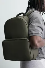 Mochila de color verde militar con textura grabada, diseño minimalista y forma redondeada. Cuenta con un compartimento principal y un bolsillo frontal, ambos con cierre de cremallera. Posee asa de mano y asas de hombro acolchadas y ajustables.