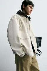 Cazadora de hombre, corte relaxed fit, color blanco hueso o crema, con acabado brillante. Presenta cuello solapa, manga larga, dos bolsillos de plastrón con solapa en la cadera y cierre frontal con botones a presión ocultos por una solapa.