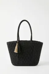 Bolso tipo tote color negro, tejido, con doble asa superior y detalle de borlas colgantes en tonos beige y negro.