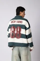 Buzo tipo polo de corte boxy, con diseño de franjas horizontales anchas en color verde oscuro y crema. Presenta cuello camisero, cierre frontal parcial y detalles de costuras desflecadas. Incluye logo bordado en el pecho.