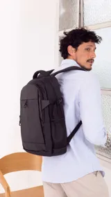 Mochila urbana de color negro con compartimento principal amplio, bolsillo frontal vertical con cierre y bolsillos laterales de red. Cuenta con correas ajustables con broches laterales, compartimento para notebook y faja trasera para sujetar a valijas.