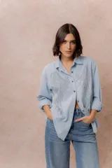 Camisa de denim liviano en tono celeste claro, con corte relajado y mangas largas. Presenta un diseño de alas bordado con strass brillante en la parte frontal y cierre mediante botones frontales.