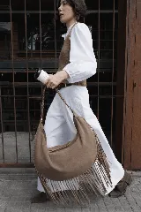 Bolso tipo hobo de gran tamaño en color marrón, confeccionado en gamuza. Presenta un diseño con flecos largos en la parte inferior, cierre superior y una correa trenzada con detalles artesanales.