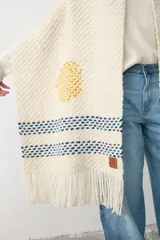 Chal tejido de punto en color crudo, con diseño artesanal que incluye un círculo amarillo y franjas horizontales en azul, con terminación de flecos en los extremos.