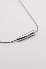 Collar de metal plateado con cadena fina estilo serpiente y un dije tubular liso y curvado en el centro. Cuenta con cierre de mosquetón y cadena de alargue.