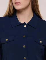 Camperita liviana de corte recto y acabado crepé color blanco, con cuello camisero clásico y cierre frontal de botones metálicos dorados. Presenta bolsillos en el pecho con solapa y mangas largas con puños abotonados.