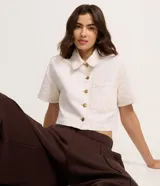 Camisa cropped de manga corta confeccionada en sarga de algodón, color crudo. Presenta cuello clásico, cierre frontal con botones metálicos, un bolsillo aplicado en el pecho y costuras pespuntadas en contraste.