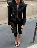 Chaqueta de estilo militar con estructura sastrera, hombros marcados y cuello mao. Presenta un diseño frontal con aplicaciones trenzadas y botones metálicos decorativos, de manga larga y corte corto.