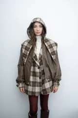 Chaqueta de cuero sintético color marrón con capucha y solapas amplias en tela escocesa a cuadros. Posee cierre frontal, puños elásticos y diseño de corte holgado.