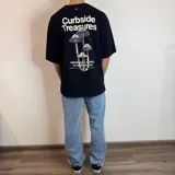 Remera negra de corte oversize con estampado gráfico en la espalda que presenta hongos en tonos violeta y verde, junto con la frase 'Curbside Treasures' y texto descriptivo.